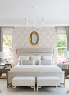 A modern bedroom featuring a large bed, two benches at the foot, matching bedside tables with lamps, a gold oval mirror above the headboard, elegant York Wallcoverings Zaria Topiary Wallpaper on the walls, and two windows with curtains allowing natural light to fill the space.