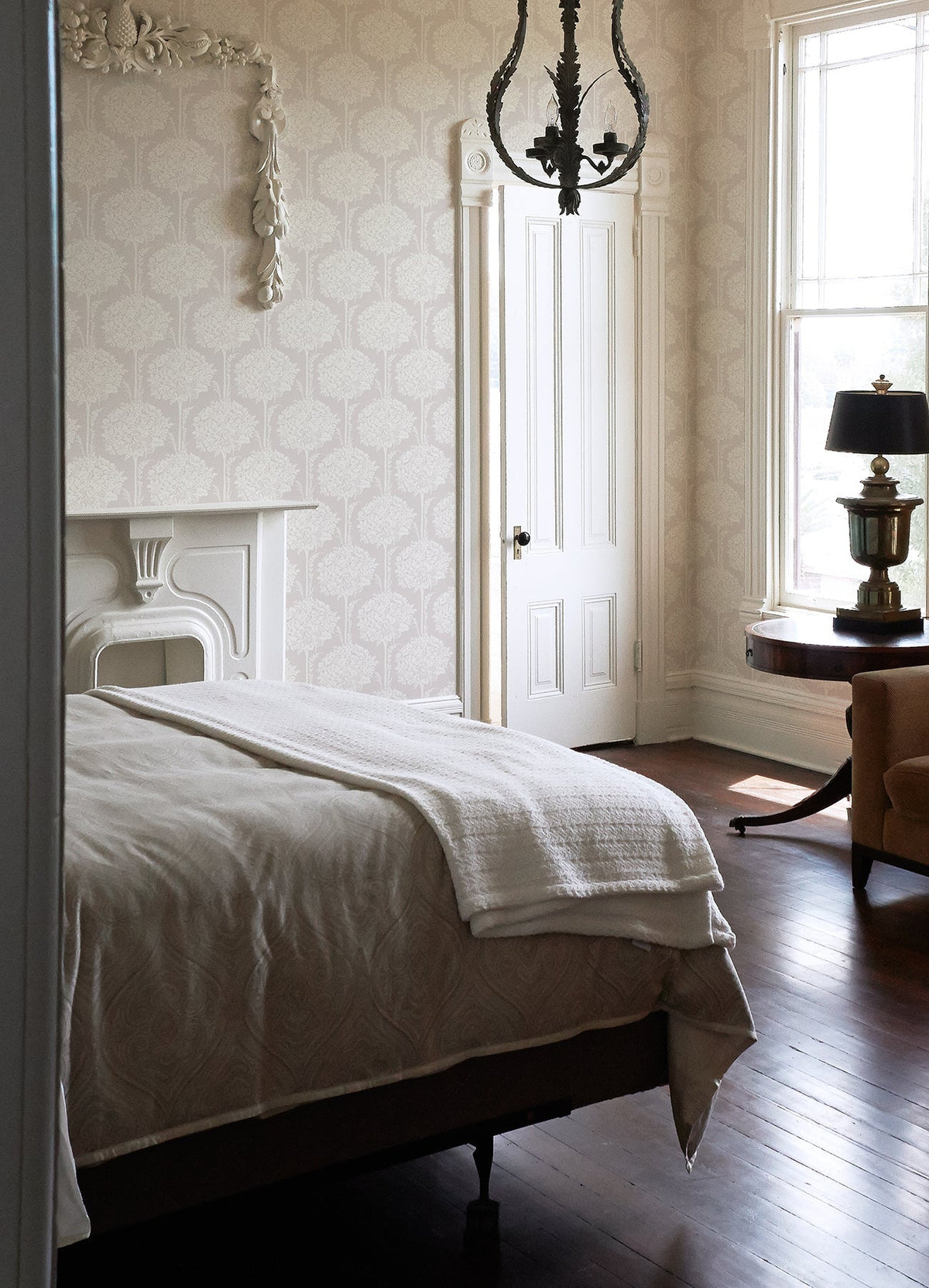 A sunlit bedroom features a neatly made bed, a white ornate fireplace, a round table with lamp, a window, chandelier, and wooden floor. Light Zaria Topiary Wallpaper by York Wallcoverings adds a soft, elegant touch.