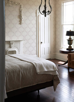 A sunlit bedroom features a neatly made bed, a white ornate fireplace, a round table with lamp, a window, chandelier, and wooden floor. Light Zaria Topiary Wallpaper by York Wallcoverings adds a soft, elegant touch.