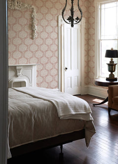 A sunlit bedroom with a cozy bed, white bedding, a fireplace, chandelier, wooden floors, and a round table with a lamp next to a large window features Zaria Topiary Wallpaper by York Wallcoverings.
