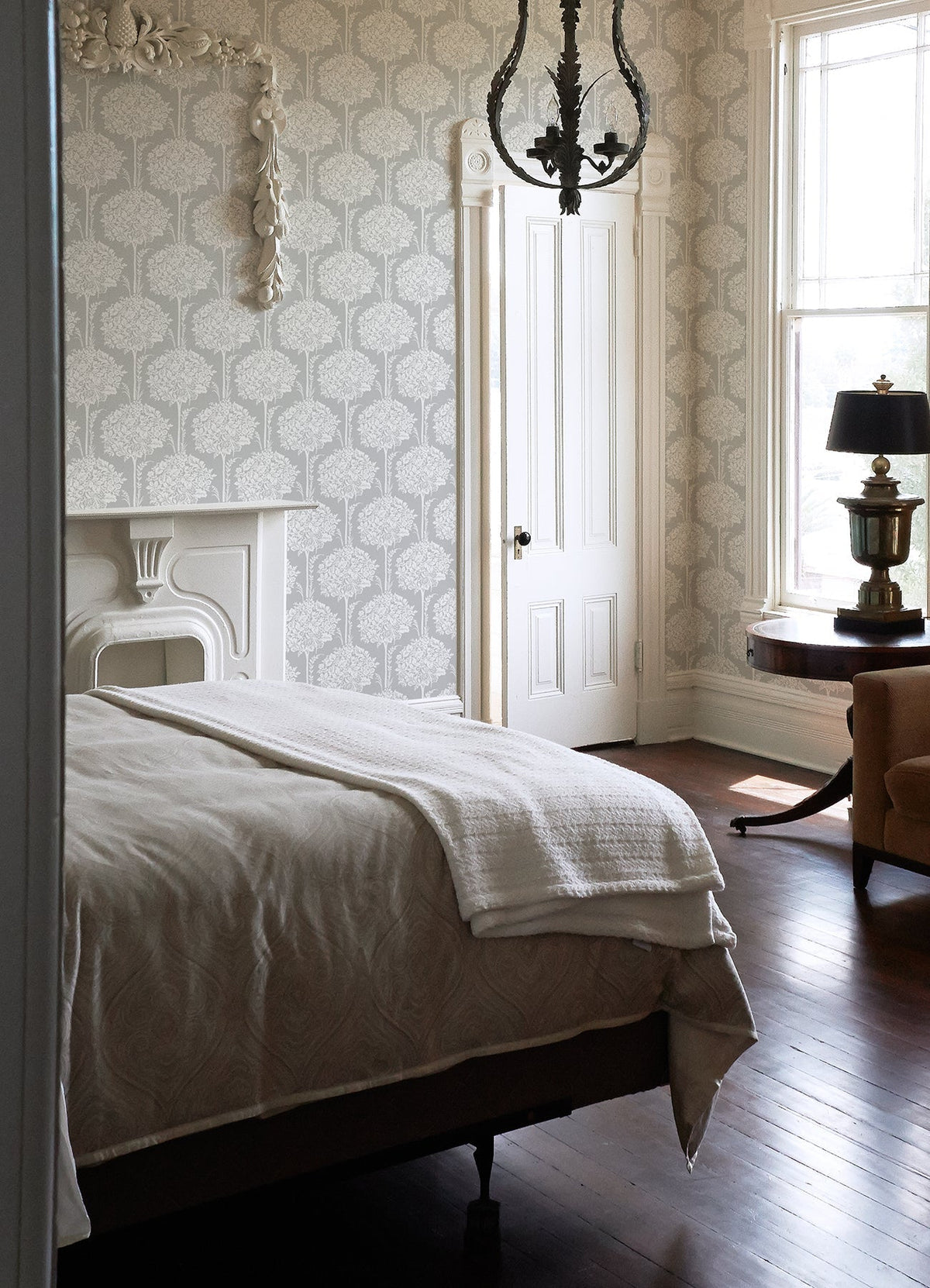 A bedroom featuring Zaria Topiary Wallpaper by York Wallcoverings, a bed with a white blanket, a white fireplace, chandelier lighting, a round table with a lamp, a tan chair, wood floors, and tall windows that fill the space with natural light.
