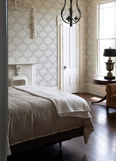 A bedroom featuring Zaria Topiary Wallpaper by York Wallcoverings, a bed with a white blanket, a white fireplace, chandelier lighting, a round table with a lamp, a tan chair, wood floors, and tall windows that fill the space with natural light.