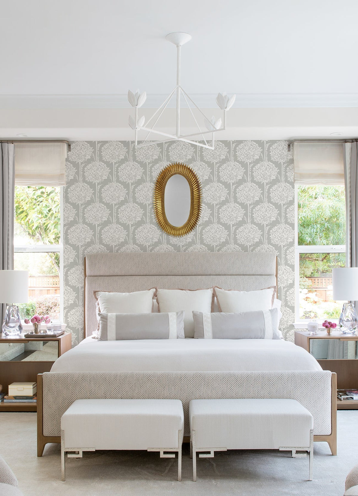 A modern bedroom featuring Zaria Topiary Wallpaper in gray by York Wallcoverings, a beige upholstered bed with white bedding, two benches, a round gold mirror, a white chandelier, and bedside tables adorned with lamps and flowers.