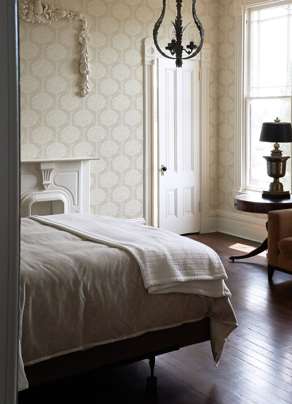 A bright bedroom featuring a neatly made bed, a white fireplace, a round table with a lamp, sunlight streaming through the window, and a beige armchair on a wooden floor—all accentuated by York Wallcoverings’ Zaria Topiary Wallpaper.