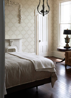 A bright bedroom featuring a neatly made bed, a white fireplace, a round table with a lamp, sunlight streaming through the window, and a beige armchair on a wooden floor—all accentuated by York Wallcoverings’ Zaria Topiary Wallpaper.