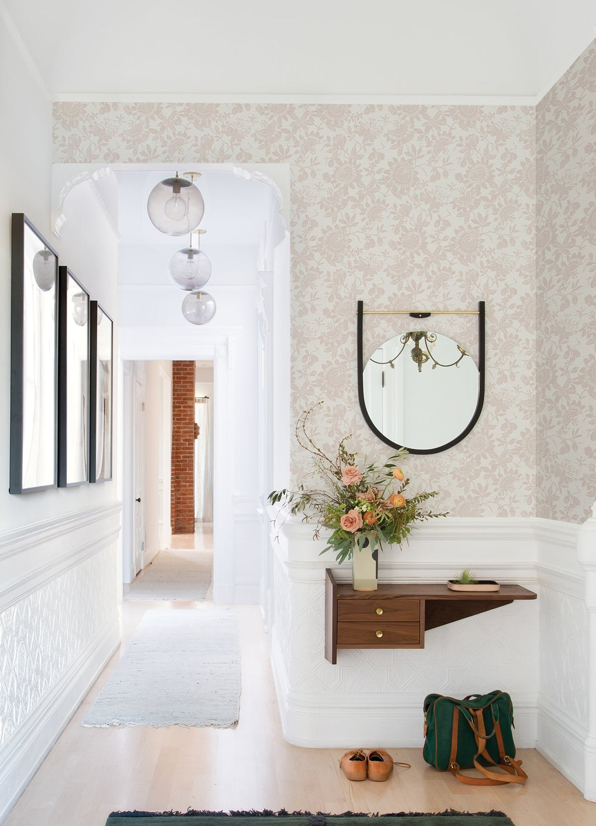 A bright hallway features Helen Floral Trail Wallpaper by York Wallcoverings, with a round mirror above a modern wood shelf displaying a vase of flowers. Shoes and a green bag rest on the floor, while three pendant lights hang from the ceiling.