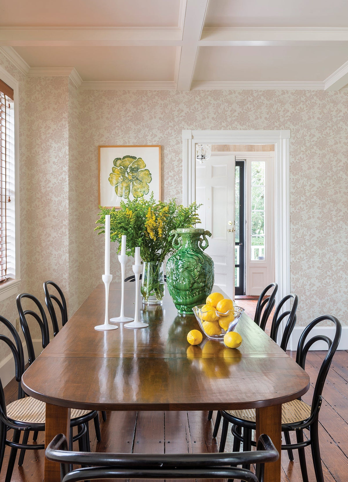 A dining room featuring Helen Floral Trail Wallpaper by York Wallcoverings, with a wooden table, black chairs, a green glass vase filled with yellow flowers, white candlesticks, and a lemon centerpiece. The space also includes wooden floors and framed art.