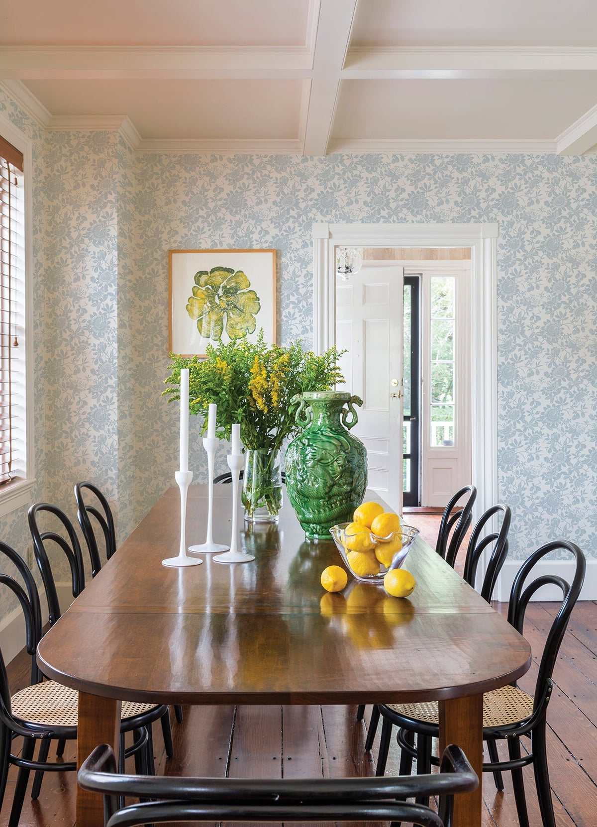 A wooden dining table with black chairs, white candlesticks, a large green vase filled with flowers, and a bowl of lemons is set in a bright room featuring York Wallcoverings’ Helen Floral Trail Wallpaper and a door leading outside.