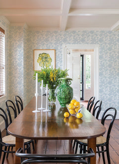 A wooden dining table with black chairs, white candlesticks, a large green vase filled with flowers, and a bowl of lemons is set in a bright room featuring York Wallcoverings’ Helen Floral Trail Wallpaper and a door leading outside.