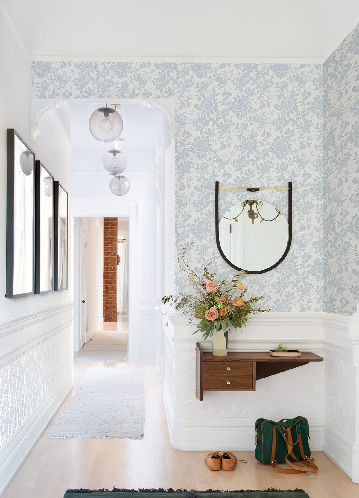 A bright hallway adorned with Helen Floral Trail Wallpaper by York Wallcoverings, featuring a round mirror above a small wooden shelf, fresh flowers, framed art, a green bag, and tan shoes on the floor. Spherical pendant lights hang from the ceiling.