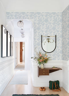 A bright hallway adorned with Helen Floral Trail Wallpaper by York Wallcoverings, featuring a round mirror above a small wooden shelf, fresh flowers, framed art, a green bag, and tan shoes on the floor. Spherical pendant lights hang from the ceiling.