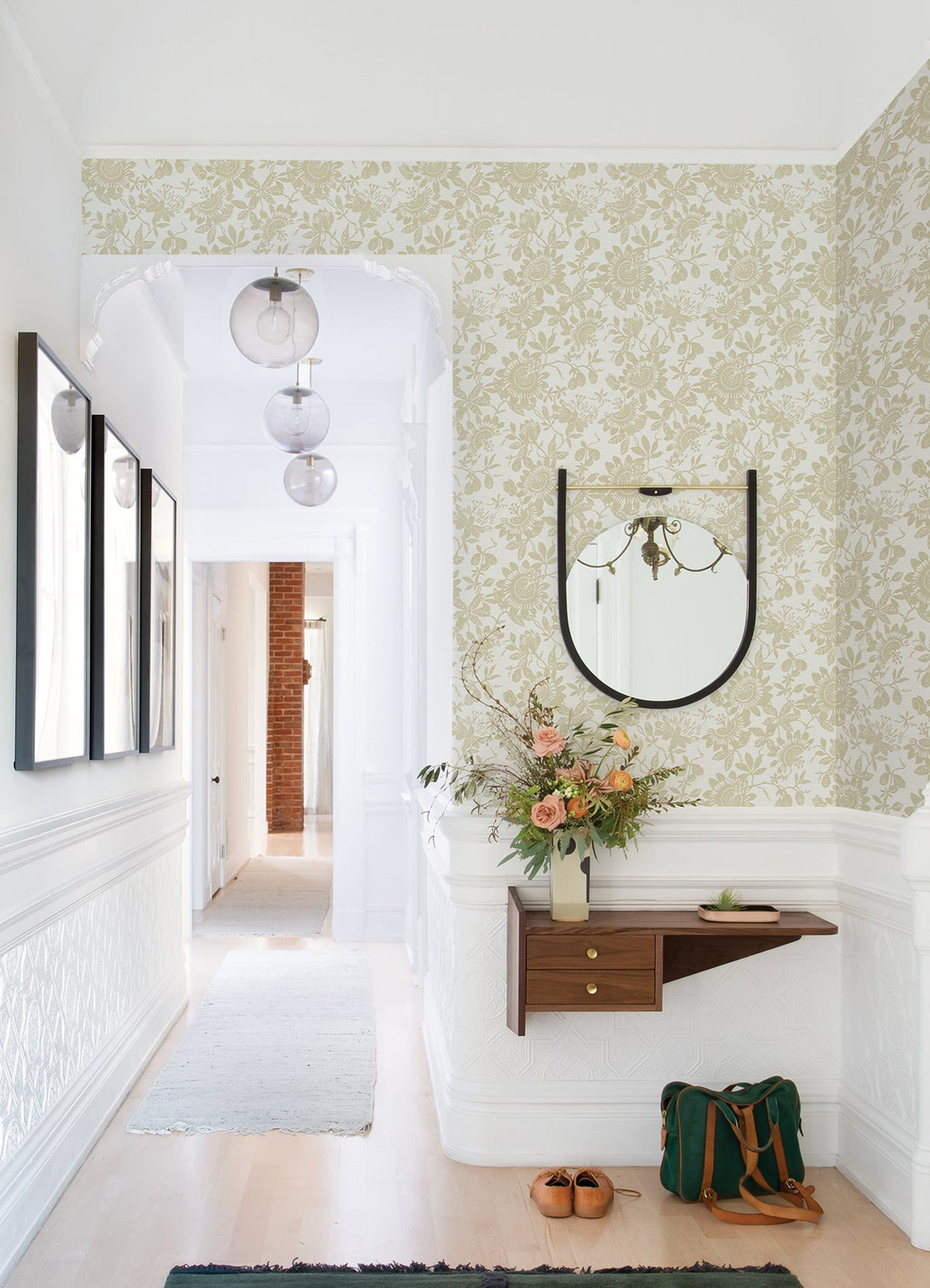 A bright hallway adorned with Helen Floral Trail Wallpaper by York Wallcoverings, featuring a round mirror above a small wooden shelf holding a vase of flowers, shoes and a green bag on the floor, and three globe pendant lights hanging from the ceiling.