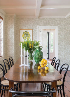 A wooden dining table with black chairs is set with white candlesticks, a green vase of yellow flowers, and a bowl of lemons. Light walls, Helen Floral Trail Wallpaper by York Wallcoverings, and a framed botanical print complete the room.