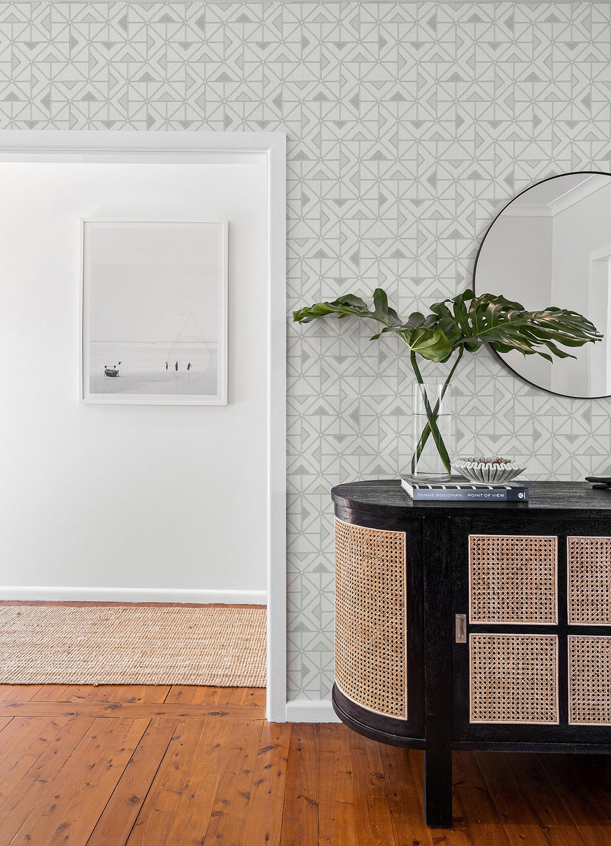 A modern entryway featuring York Wallcoverings’ Jekyl Triangles Wallpaper, a round mirror, a black and rattan console table, green leaves in a vase, books, and wall art in an adjacent room with wooden floors.
