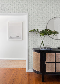 A modern entryway featuring York Wallcoverings’ Jekyl Triangles Wallpaper, a round mirror, a black and rattan console table, green leaves in a vase, books, and wall art in an adjacent room with wooden floors.