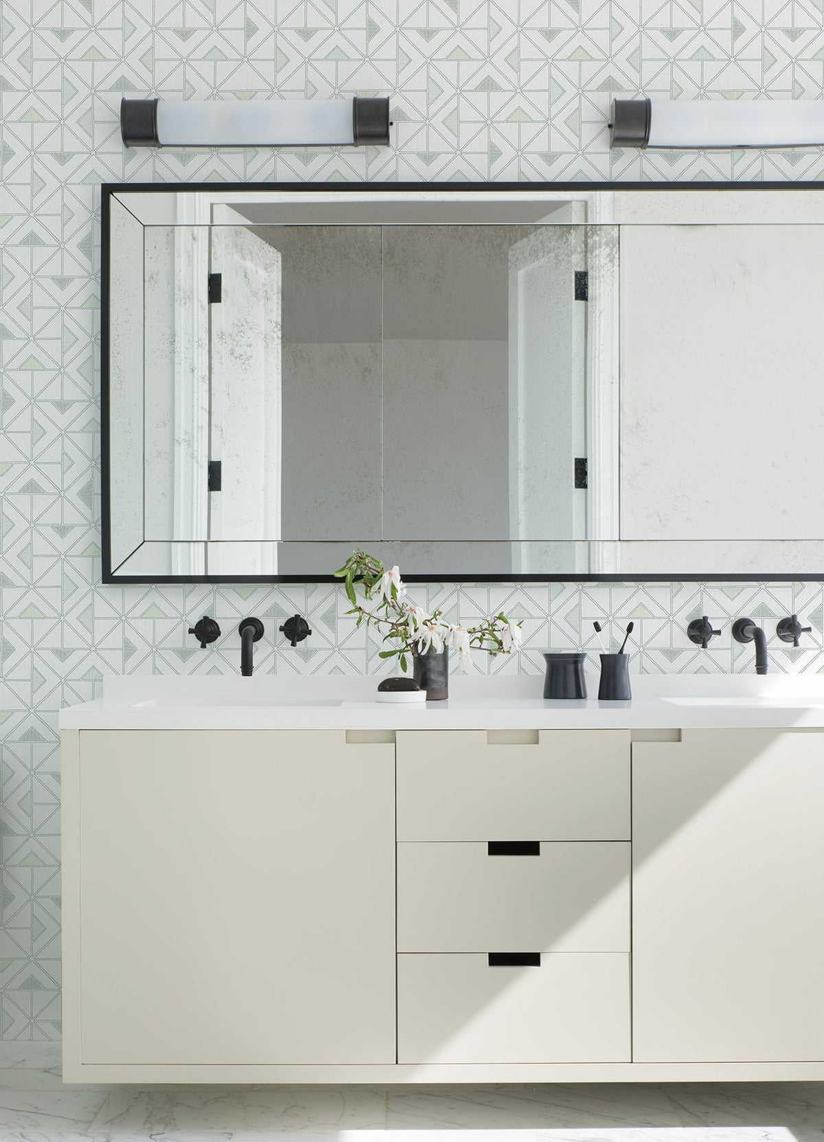 Modern bathroom featuring a large mirror, Jekyl Triangles Wallpaper by York Wallcoverings on the walls, a white vanity with black hardware, two wall lights above, and minimal accessories like a vase with green branches.