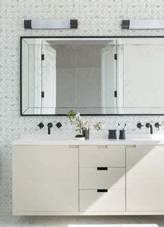 Modern bathroom featuring a large mirror, Jekyl Triangles Wallpaper by York Wallcoverings on the walls, a white vanity with black hardware, two wall lights above, and minimal accessories like a vase with green branches.