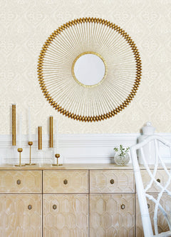 A room features Scout Floral Ogee Wallpaper by York Wallcoverings as a stylish backdrop. A round gold decorative mirror hangs above a light wood cabinet with geometric patterns, which is adorned with gold and glass candle holders and a small vase with greenery; part of a white chair is also visible.