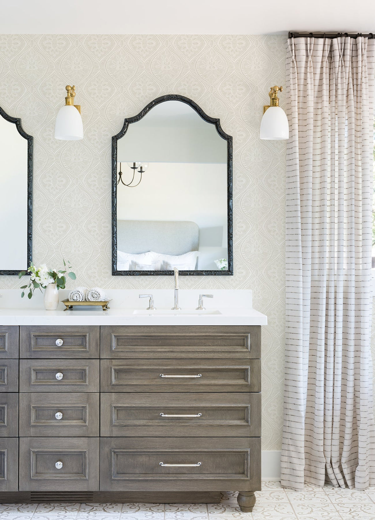 Bright bathroom featuring Scout Floral Ogee Wallpaper by York Wallcoverings, double mirrors above a wood vanity with drawers, two white light sconces, a vase with flowers, and floor-length striped curtains beside the sink.