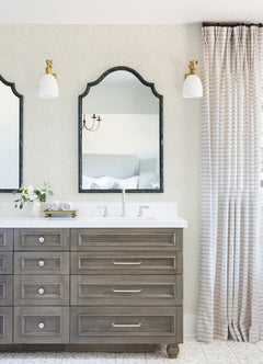 Bright bathroom featuring Scout Floral Ogee Wallpaper by York Wallcoverings, double mirrors above a wood vanity with drawers, two white light sconces, a vase with flowers, and floor-length striped curtains beside the sink.
