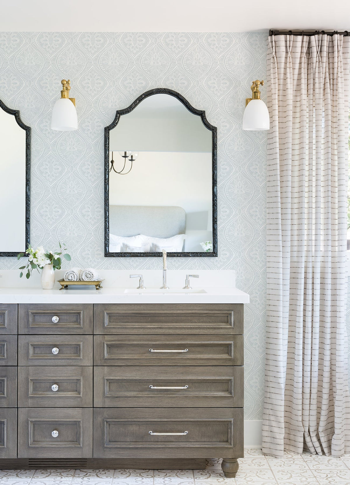 A bathroom featuring a double sink vanity, two mirrors, sconce lights, and striped curtains is adorned with York Wallcoverings’ Scout Floral Ogee Wallpaper. The countertop is decorated with a small vase of flowers and a clock.