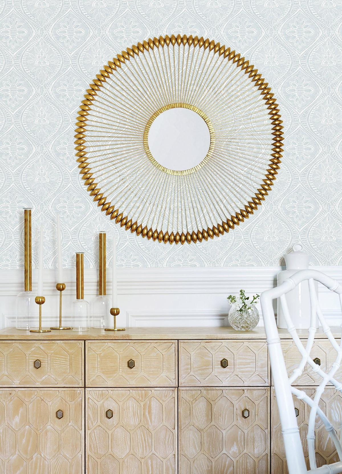 The wall behind a light wood cabinet is adorned with Scout Floral Ogee Wallpaper by York Wallcoverings, while glass candle holders and a small vase sit on top of the cabinet. A white chair is partially visible in the foreground.