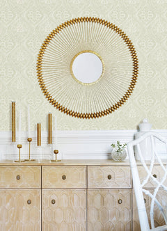 A round gold decorative mirror hangs on a wall adorned with Scout Floral Ogee Wallpaper by York Wallcoverings above a light wood cabinet. The cabinet displays gold-accented candleholders and a small vase with greenery, while a white chair is partially visible.