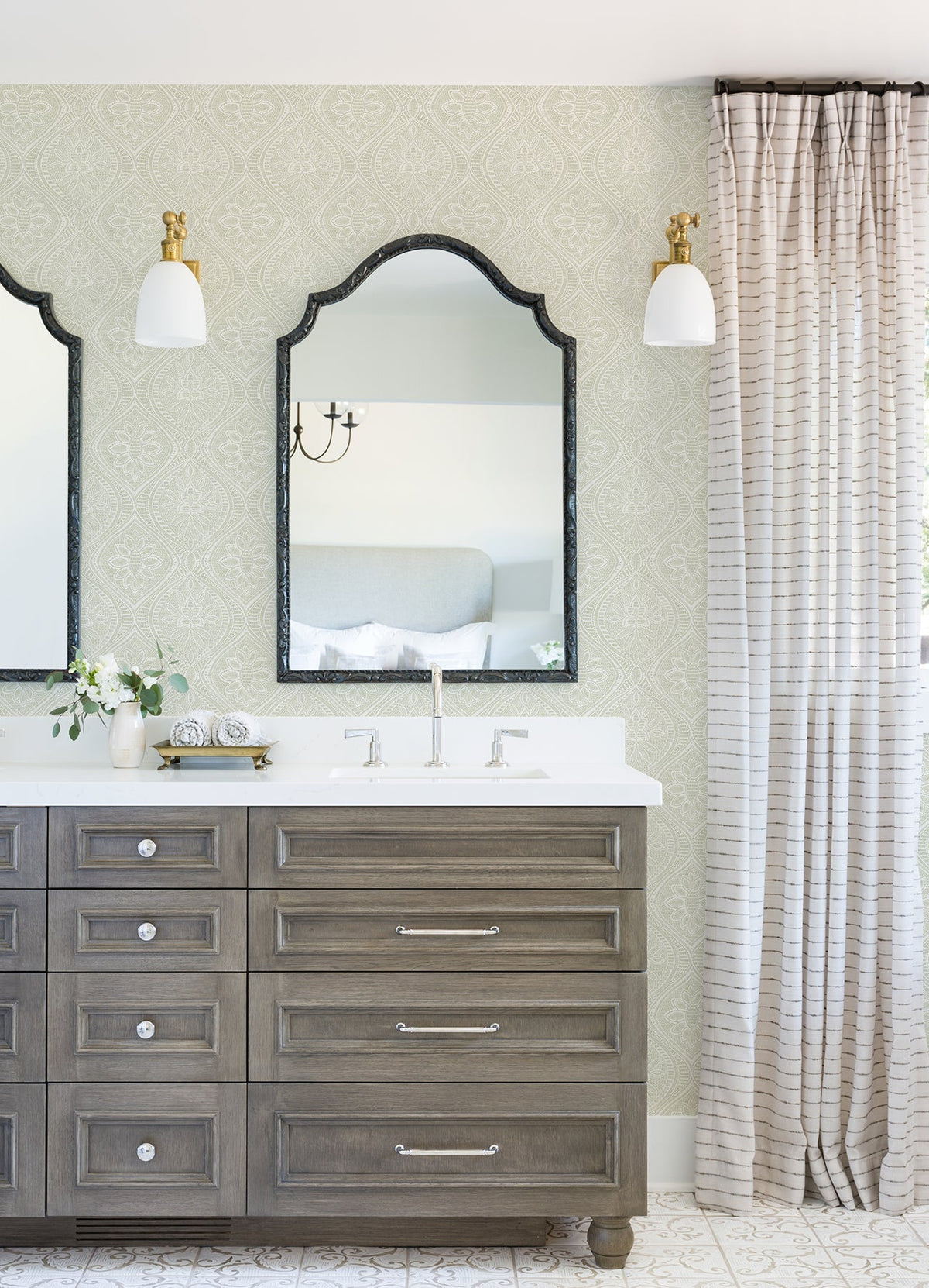 A bathroom vanity with two mirrors, double sinks, light fixtures above each mirror, gray drawers, a small vase with flowers, and Scout Floral Ogee Wallpaper by York Wallcoverings adorning the walls beside the vanity.