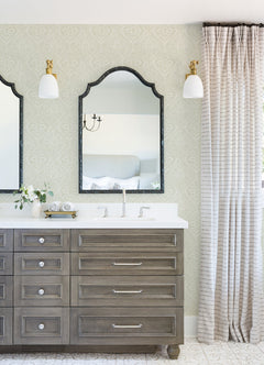 A bathroom vanity with two mirrors, double sinks, light fixtures above each mirror, gray drawers, a small vase with flowers, and Scout Floral Ogee Wallpaper by York Wallcoverings adorning the walls beside the vanity.