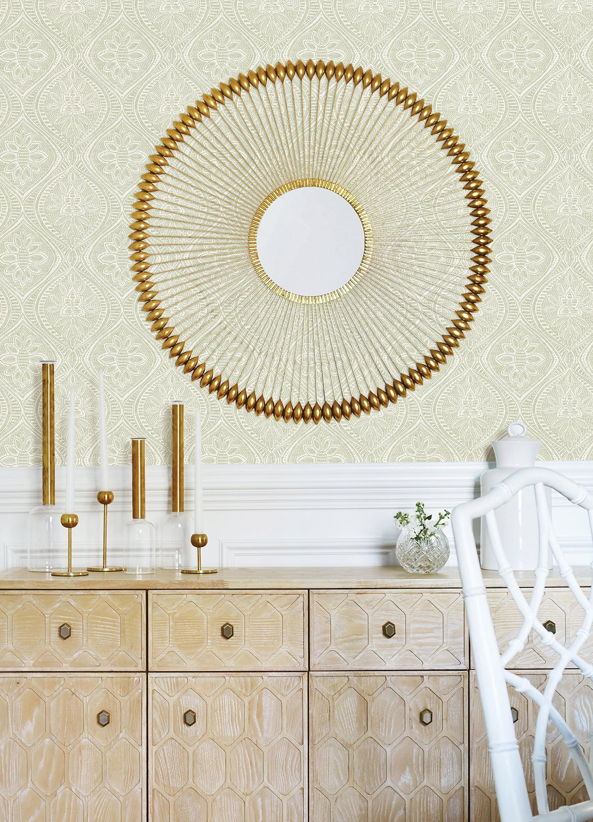 A gold sunburst mirror hangs above a light wood cabinet with geometric patterns, which displays gold and glass candle holders and a vase filled with greenery. In the foreground sits a white chair, while the walls are adorned with Scout Floral Ogee Wallpaper by York Wallcoverings in the background.