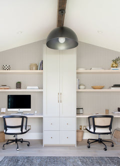 A modern home office features Glynn Chevron Wallpaper by York Wallcoverings, two desks, black and white chairs, shelves with decor, a central cabinet, a desktop computer, and a large black dome pendant light hanging from a beamed ceiling.