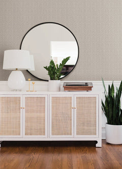A white sideboard with wicker doors holds a white lamp, two gold candlesticks, potted plants, and a record player. A round mirror hangs above. The floor is wooden, and the wall is adorned with Glynn Chevron Wallpaper by York Wallcoverings, featuring a subtle herringbone pattern.