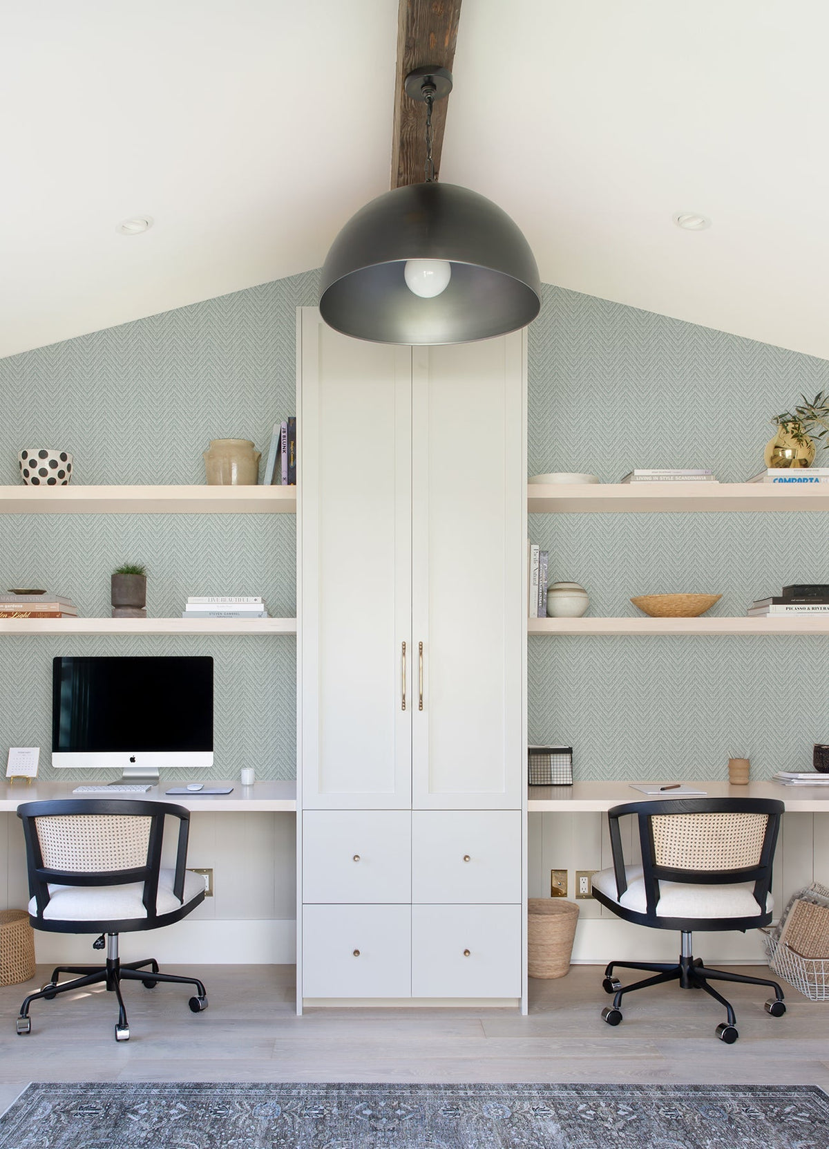 Transformed Sentence: Modern home office featuring two desks, black office chairs, a large overhead light fixture, built-in white shelves displaying books and decor, a desktop computer, and neutral minimalist accents—all brought together by Glynn Chevron Wallpaper from York Wallcoverings.