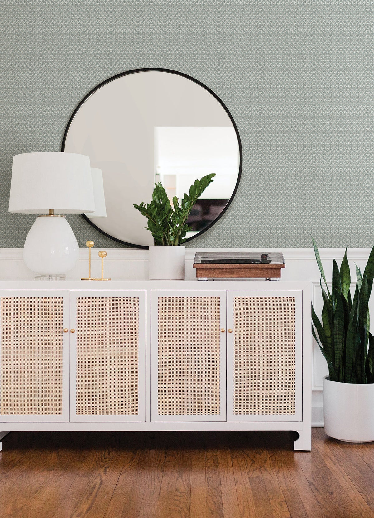 A modern sideboard with cane doors holds a white lamp, two gold candle holders, potted plants, and a record player. A large round mirror hangs above, reflecting the room. Wooden floors complement the walls covered in Glynn Chevron Wallpaper by York Wallcoverings.