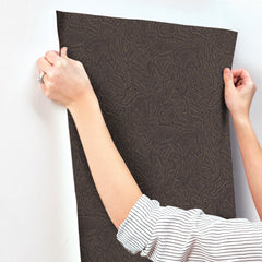 A person in a striped shirt uses both hands to smooth or position the Hollenback Dotted Abstract Wallpaper by York Wallcoverings against a white wall.