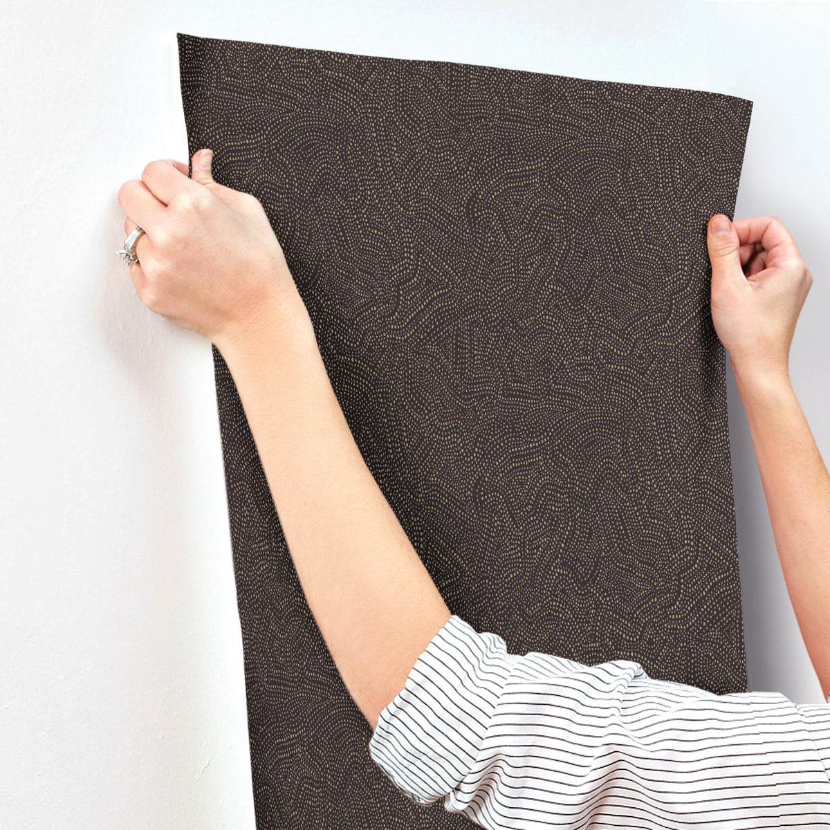 A person in a striped shirt holds up York Wallcoverings’ Hollenback Dotted Abstract Wallpaper against a white wall, preparing to apply it.