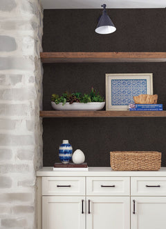 White cabinets with black handles stand beneath two wooden shelves displaying plants, a blue vase, wicker basket, framed tile print, and a bowl—all set against a dark wall adorned with York Wallcoverings’ Hollenback Dotted Abstract Wallpaper and highlighted by an overhead sconce light.