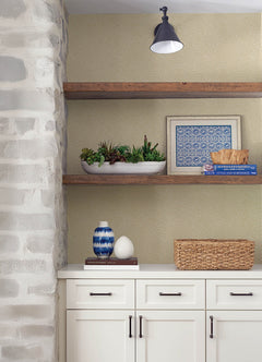 White cabinets with black handles are set beneath floating wooden shelves, which display a planter of succulents, books, framed art, a basket, and a blue-and-white vase. The backdrop features York Wallcoverings’ Hollenback Dotted Abstract Wallpaper, highlighted by a wall sconce.