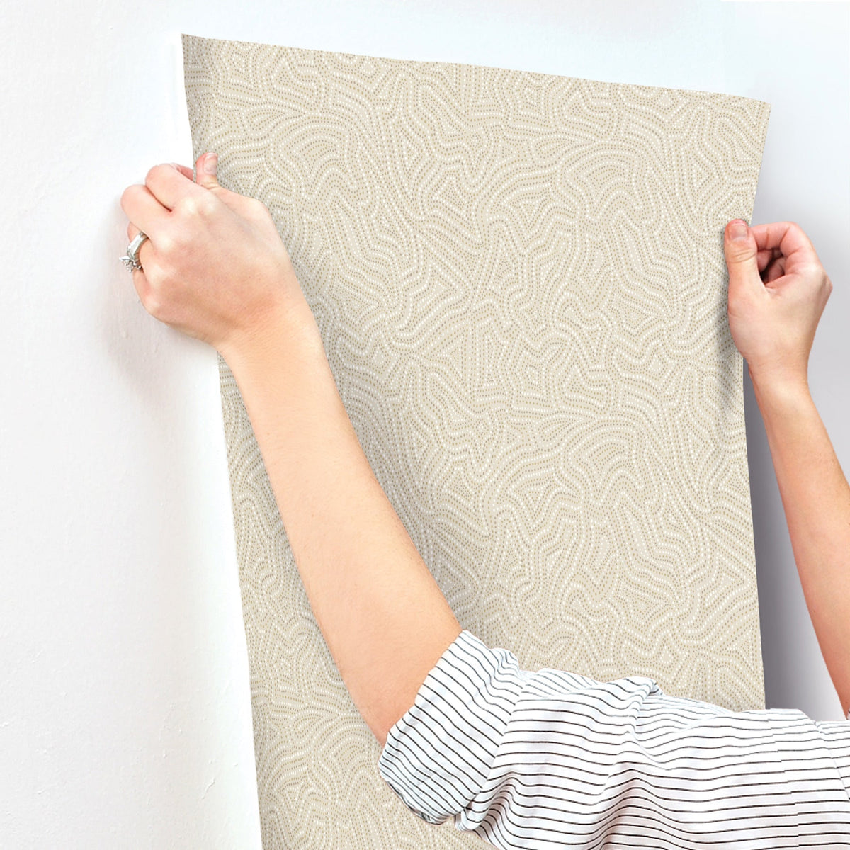 A person with a striped sleeve uses both hands to apply Hollenback Dotted Abstract Wallpaper by York Wallcoverings to a white wall.