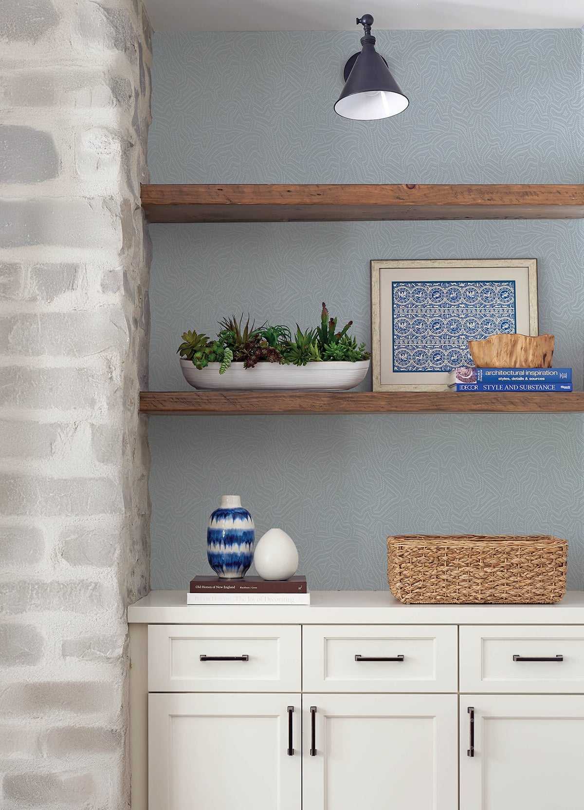 White shelves adorned with potted succulents, blue-patterned art, a decorative bowl, vase, and wicker basket are complemented by a stone wall and a white cabinet with black handles. The area also features the Hollenback Dotted Abstract Wallpaper by York Wallcoverings as its gray patterned wall.