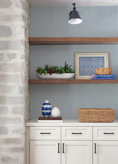 White shelves adorned with potted succulents, blue-patterned art, a decorative bowl, vase, and wicker basket are complemented by a stone wall and a white cabinet with black handles. The area also features the Hollenback Dotted Abstract Wallpaper by York Wallcoverings as its gray patterned wall.