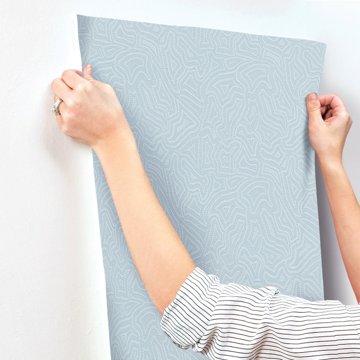 A person wearing a striped shirt is aligning a sheet of Hollenback Dotted Abstract Wallpaper by York Wallcoverings, featuring a light blue pattern, on a white wall.