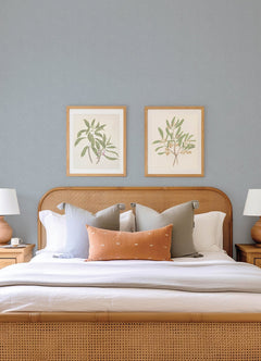 A neatly made bed with neutral pillows and a tan headboard is framed by two wooden nightstands with lamps. Two botanical prints hang on the Hollenback Dotted Abstract Wallpaper by York Wallcoverings above the bed.
