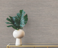 A white vase holding monstera leaves sits on a narrow metal table against gray Winn Faux Grasscloth Wallpaper by York Wallcoverings.