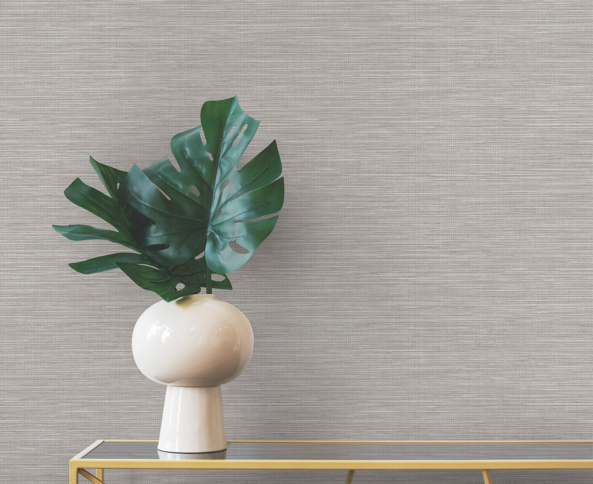 A white vase containing large green monstera leaves sits on a gold console table against neutral-toned Winn Faux Grasscloth Wallpaper by York Wallcoverings.