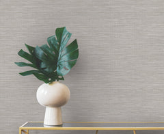 A white vase containing large green monstera leaves sits on a gold console table against neutral-toned Winn Faux Grasscloth Wallpaper by York Wallcoverings.