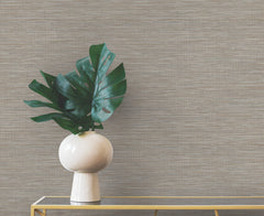A white vase containing large green monstera leaves sits on a gold console table against a neutral-toned Winn Faux Grasscloth Wallpaper by York Wallcoverings.