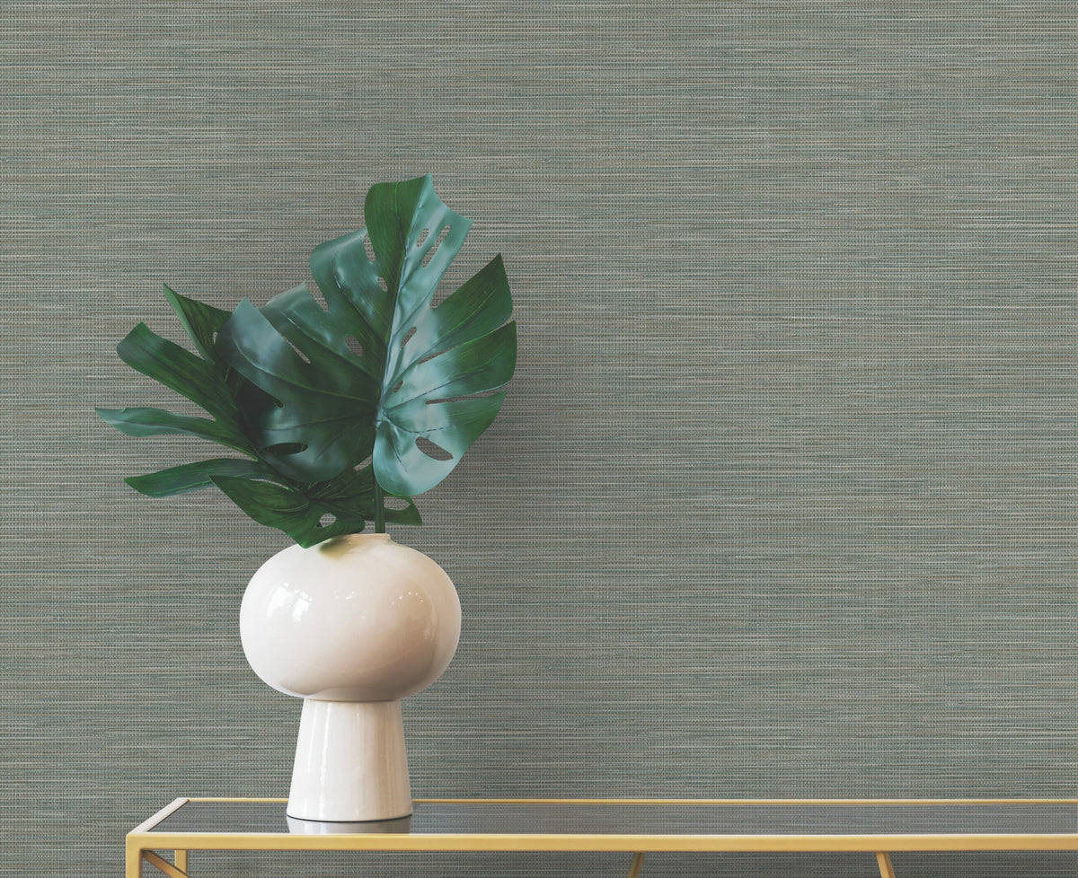 A monstera plant in a white vase sits on a gold-framed table in front of gray Winn Faux Grasscloth Wallpaper by York Wallcoverings.