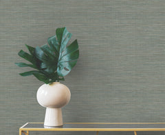 A monstera plant in a white vase sits on a gold-framed table in front of gray Winn Faux Grasscloth Wallpaper by York Wallcoverings.