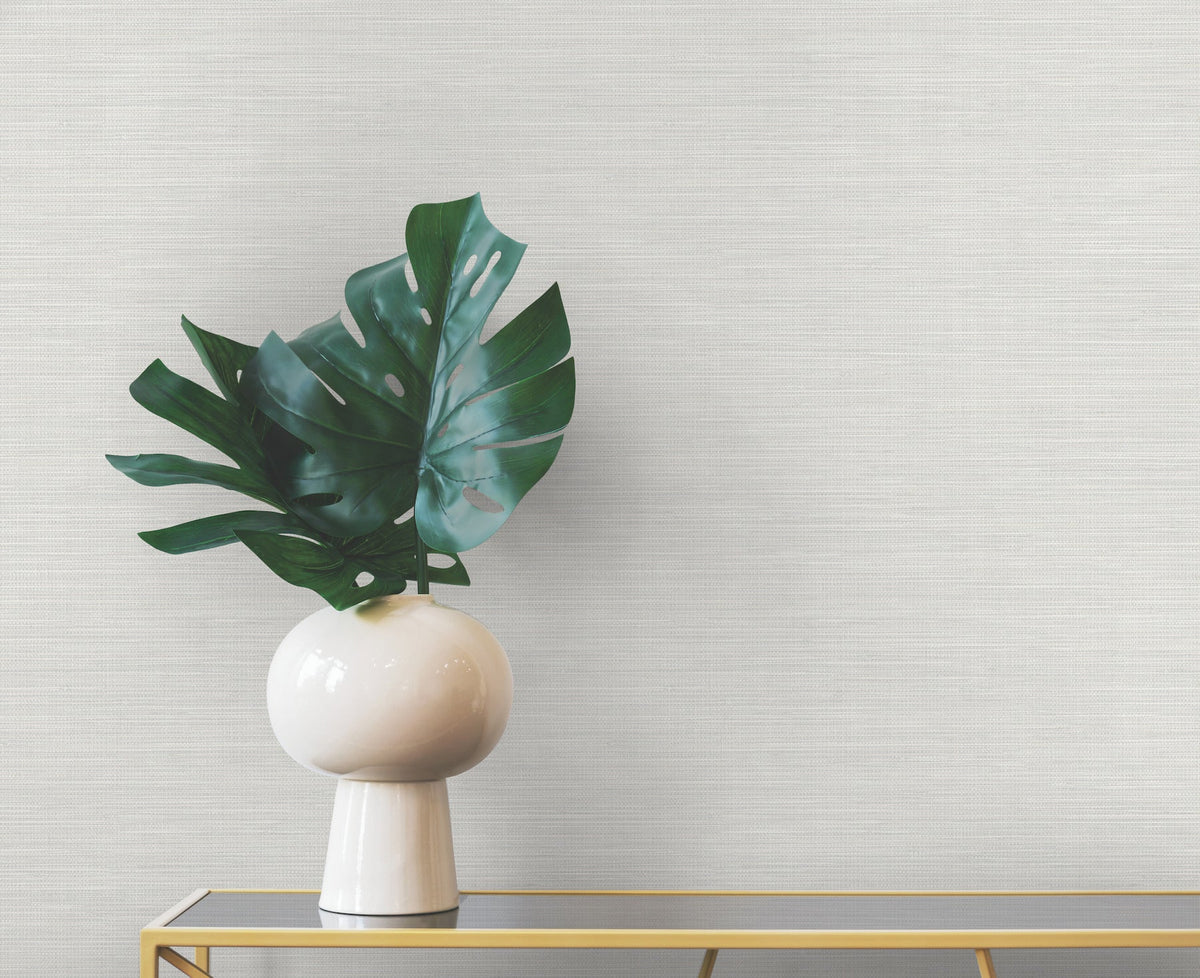 A vase containing large green monstera leaves sits on a narrow table against a wall with textured light gray Winn Faux Grasscloth Wallpaper.